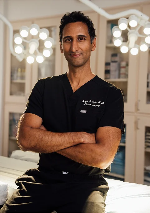 A professional portrait of Dr. Surjit Rai in black surgical scrubs with "Plastic Surgery" embroidered on the chest. He stands with arms crossed in a brightly lit clinical room with surgical lights in the background.