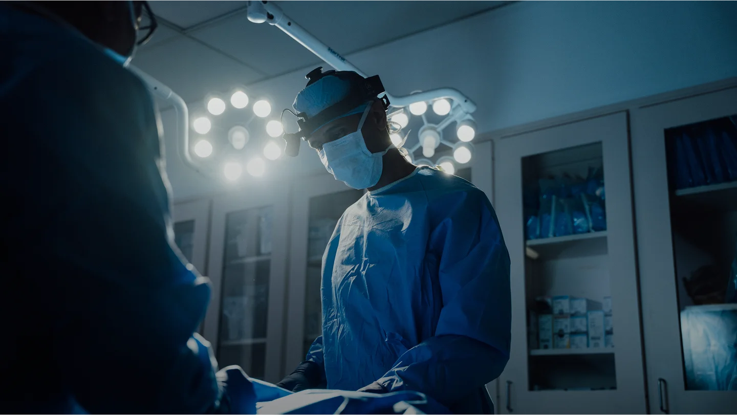 A dramatic, low-light shot of a surgeon (Dr. Rai) in a sterile operating room. He is wearing blue surgical scrubs, a mask, and a surgical headlamp, focused intently on a procedure under bright overhead surgical lights.