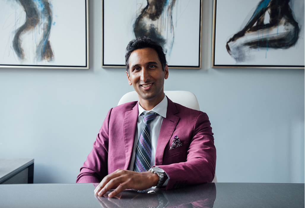 A professional portrait of Dr. Surjit Rai sitting at a sleek desk. He is wearing a mauve-colored suit jacket, a striped tie, and a floral pocket square. Three abstract pieces of art depicting the human form hang on the wall behind him.