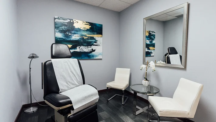 A clean and modern examination room featuring a black adjustable medical chair covered with a sterile white sheet. The room is decorated with a large piece of abstract teal and gold art, two white guest chairs, and a sleek glass side table with a white orchid.