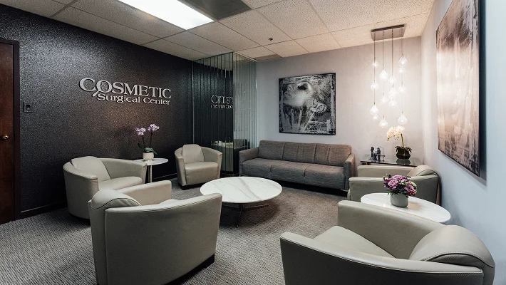 The main reception and waiting lounge of the Cosmetic Surgical Center. The room features plush grey seating, marble-topped tables, and a large "Cosmetic Surgical Center" logo on a shimmering black feature wall. Modern teardrop-shaped pendant lights hang in the corner.