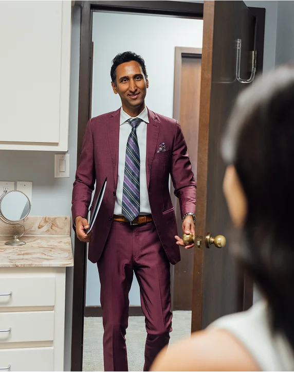Dr. Surjit Rai, carrying a black patient folder, smiles as he opens a door to enter an examination room. He is dressed in a professional purple suit and striped tie.