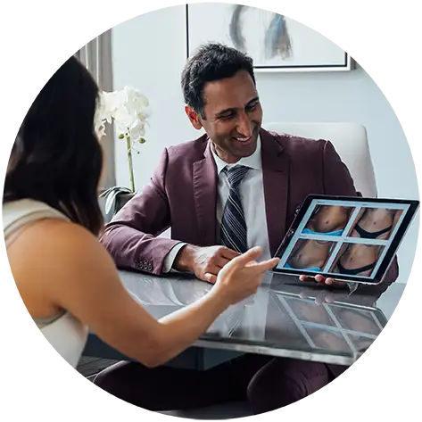 Dr. Surjit Rai, wearing a professional mauve suit, conducts a consultation with a female patient. He is smiling as he uses a tablet to show her "before and after" photos of a body contouring procedure.