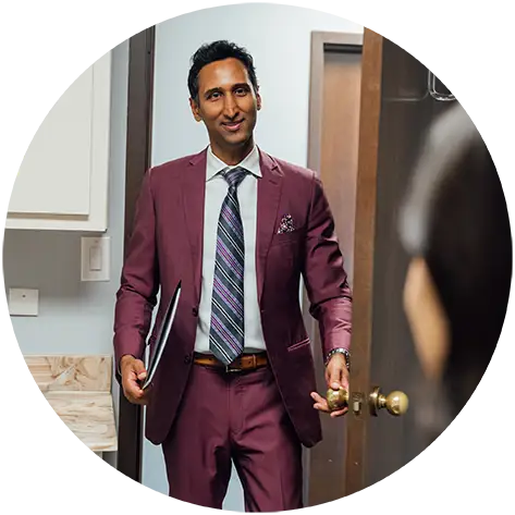 Dr. Surjit Rai, carrying a black patient folder, smiles as he opens a door to enter an examination room. He is dressed in a professional purple suit and striped tie.