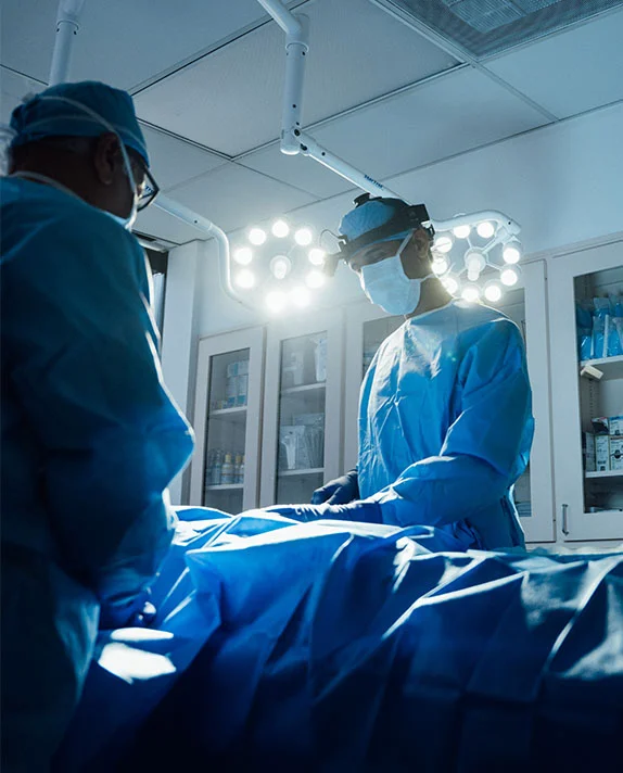 A professional surgical scene showing Dr. Rai in blue scrubs, masks, and headlamps performing a procedure under bright operating room lights.