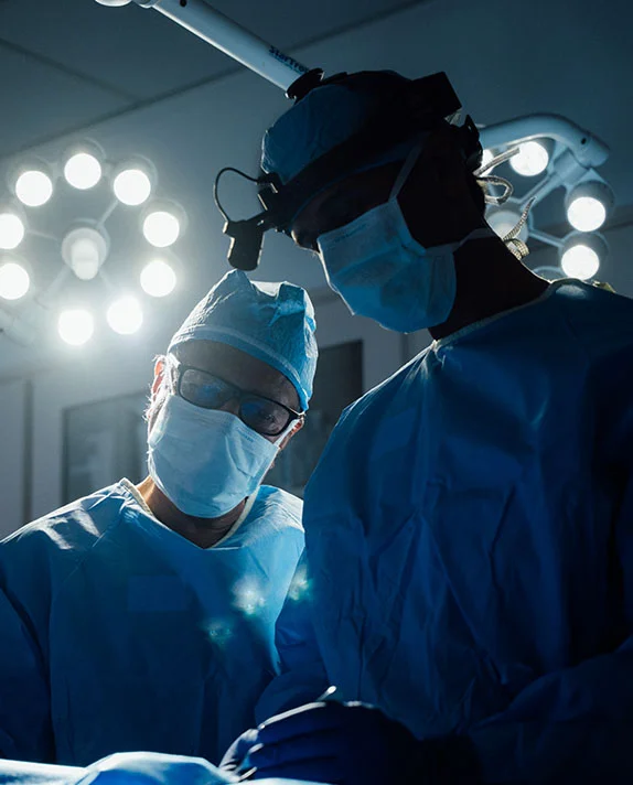 An wide-angle view of a surgical team, including Dr. Rai, working in a darkened operating room under a bright circular surgical light. A large window shows a blue sky in the background.