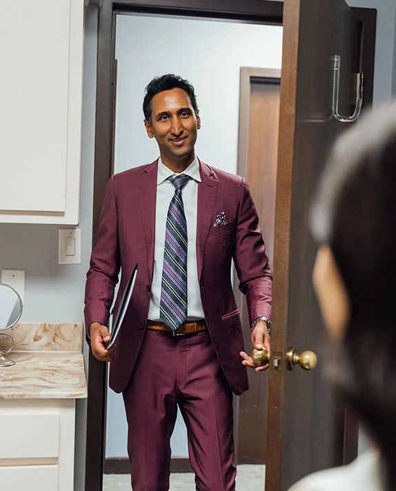 Dr. Surjit Rai, carrying a black patient folder, smiles as he opens a door to enter an examination room. He is dressed in a professional purple suit and striped tie.
