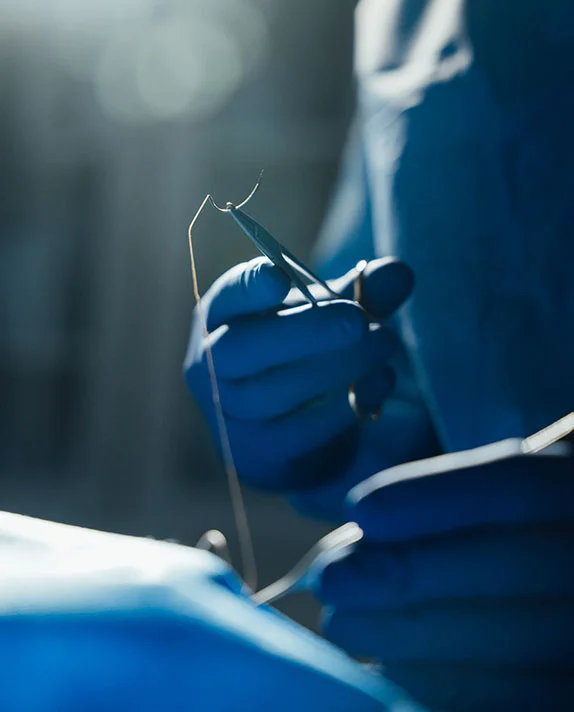 A close-up, artistic shot of a surgeon’s gloved hands in a blue-tinted operating room. The surgeon is holding a needle driver and surgical suture, representing the precision of a surgical procedure.
